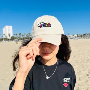 May include: A beige baseball cap with the word "PEACE" embroidered in colorful letters. The cap is worn with a black t-shirt featuring the text "JANYA SOUL" and a red heart graphic. A silver necklace and bracelet are also visible.