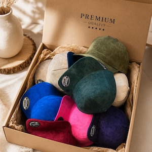 May include: A collection of multi-coloured suede-textured snapback caps packed inside a cardboard box labelled PREMIUM QUALITY with five stars. The caps feature various colours including olive green, dark green, blue, pink, purple, and beige.