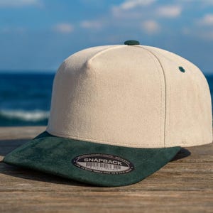 Op de afbeelding: Een beige en donkergroene snapback cap met een platte klep. De cap heeft een beige kroon en een donkergroene klep. Het heeft een kleine groene knop bovenop en een "SNAPBACK" label. De achtergrond toont een houten oppervlak en de zee.