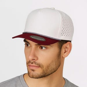 May include: A white baseball cap with a burgundy peak and a perforated side panel. The cap has a decorative rope detail along the peak's edge. The hat is designed with a flat peak and a structured crown.