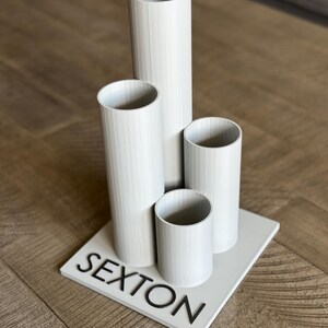 May include: A white decorative piece featuring four cylindrical tubes of varying heights, arranged on a square base. The word "SEXTON" is displayed in black, block letters on the base. The piece is set against a wooden surface.