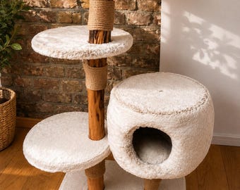 Op de afbeelding: Een krabpaal met meerdere niveaus, voorzien van drie ronde plateaus en een gesloten ronde slaaphol, bekleed met crèmekleurige pluche stof. De constructie wordt ondersteund door natuurlijke houten palen omwikkeld met dik, beige sisaltouw.