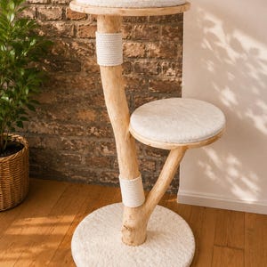 Puede incluir: Un árbol para gatos con una estructura de tronco de madera de color claro natural, dos plataformas circulares y una base redonda. Cada plataforma y la base están cubiertas con una tela blanca suave. Dos secciones del tronco están envueltas en cuerda blanca.