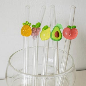 Puede incluir: Pajitas de vidrio transparente con adornos de frutas coloridas: piña, uvas, limón, aguacate y melocotón. Las pajitas se muestran en un cuenco de vidrio transparente, ideales para bebidas.