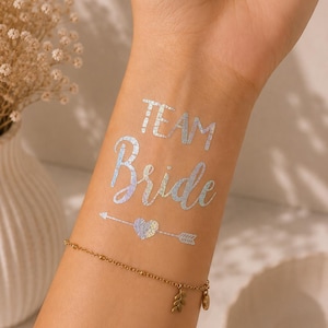 Op de afbeelding: Een tijdelijke tatoeage op de huid met de iriserende zilveren tekst "TEAM Bride" boven een klein hartje dat door een pijl wordt doorboord. Het ontwerp heeft een metallic, holografische afwerking die licht reflecteert, boven een dunne gouden armband.