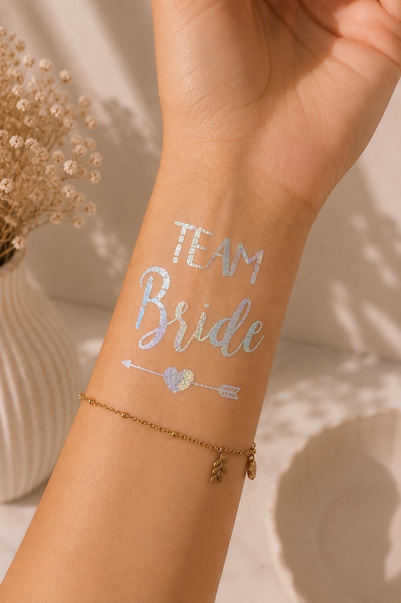 Op de afbeelding: Een tijdelijke tatoeage op de huid met de iriserende zilveren tekst "TEAM Bride" boven een klein hartje dat door een pijl wordt doorboord. Het ontwerp heeft een metallic, holografische afwerking die licht reflecteert, boven een dunne gouden armband.