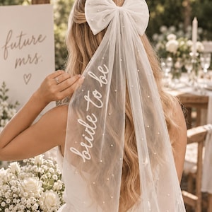 Könnte beinhalten: Ein weißer Tüll-Haarschleier mit einer großen Schleife, verziert mit kleinen, verstreuten weißen Perlen und dem Schriftzug bride to be in weißer Schreibschrift. Der Schleier besteht aus transparentem Stoff und fällt locker über den Rücken.