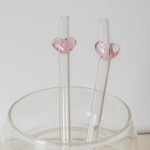 Op de afbeelding: Twee heldere glazen rietjes met roze hartvormige versieringen liggen in een heldere glazen kom. De rietjes zijn cilindrisch en de hartversieringen zijn zachtroze. De kom is rond en transparant.