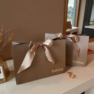 Op de afbeelding: Twee taupekleurige cadeauzakken met satijnen strikken. Op de ene tas staat "Sabine", op de andere "Melina". De tassen hebben touwhandvatten en staan op een wit oppervlak. Een gouden ring met twee bollen ligt ervoor.