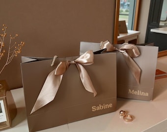 Può includere: Due sacchetti regalo color talpa con fiocchi di nastro di raso. Su una borsa è stampato "Sabine", sull'altra "Melina". Le borse hanno manici in corda e sono su una superficie bianca. Davanti c'è un anello d'oro con due sfere.