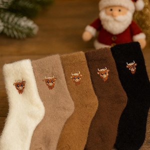 Peut inclure: Une collection de cinq paires de chaussettes duveteuses, crème, beige, marron clair, marron foncé et noires. Chaque chaussette présente un motif de taureau brodé. Un Père Noël et un petit sapin de Noël sont visibles en arrière-plan.