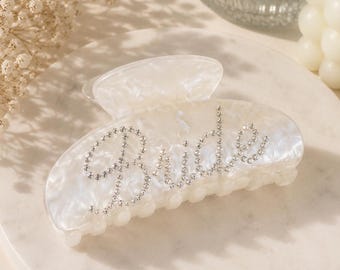 Op de afbeelding: Een witte, gemarmerde haarklem met het woord Bride in kleine, heldere, fonkelende strass-steentjes. Het accessoire heeft een gebogen, semi-transparant ontwerp met een veermechanisme en in elkaar grijpende tanden voor een stevige grip.