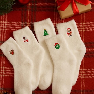 Puede incluir: Cinco calcetines navideños blancos y afelpados con diseños bordados: un reno, un muñeco de nieve, un árbol de Navidad, una corona y Papá Noel. Dispuestos sobre una superficie a cuadros rojos con una caja de regalo y una piña.