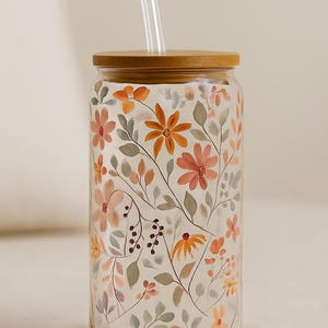 Op de afbeelding: Een heldere glazen beker met een bamboe deksel en een helder rietje. De beker is versierd met een bloemenpatroon in tinten oranje, perzik en groen. Het ontwerp bevat verschillende bloemen en bladeren.