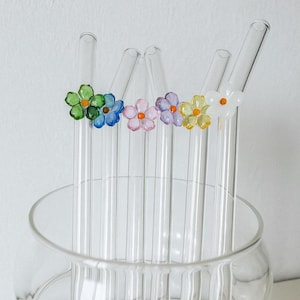 Op de afbeelding: Transparante glazen rietjes met kleurrijke bloemaccenten. De bloemen zijn groen, blauw, roze, paars, geel en wit. De rietjes worden getoond in een transparante glazen kom. De achtergrond is een effen witte muur.