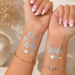 Op de afbeelding: Twee polsen met iriserende tijdelijke tatoeages. De ene pols toont de tekst "TEAM Bride" boven een pijl- en hartafbeelding. De andere pols toont de tekst "Bride" boven een pijl- en hartafbeelding. Beide tatoeages hebben een reflecterende, holografische afwerking.