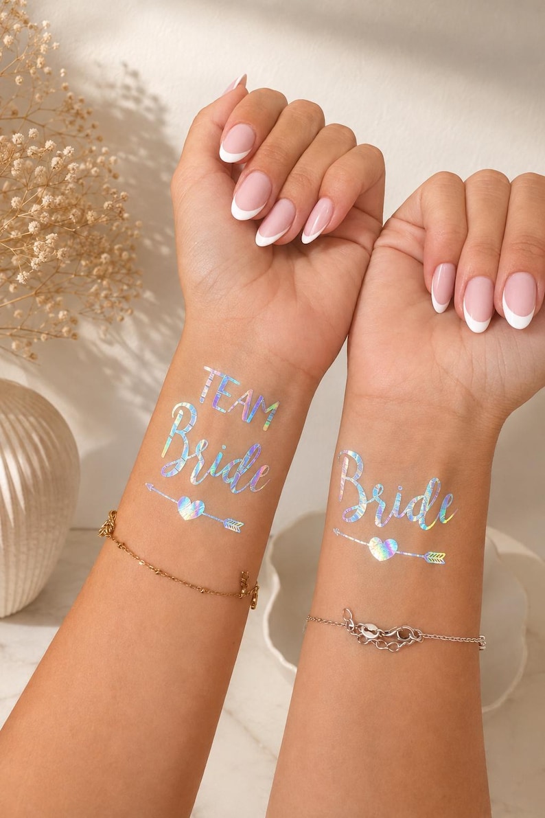 Op de afbeelding: Twee polsen met iriserende tijdelijke tatoeages. De ene pols toont de tekst "TEAM Bride" boven een pijl- en hartafbeelding. De andere pols toont de tekst "Bride" boven een pijl- en hartafbeelding. Beide tatoeages hebben een reflecterende, holografische afwerking.