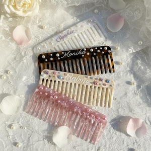 May include: Four personalised hair combs in white, tortoiseshell, cream, and pink. Each comb is decorated with pearls and has a name written on it: Sophie, Monika, Sabine, and Salome. The combs are on a white lace surface with flower petals.