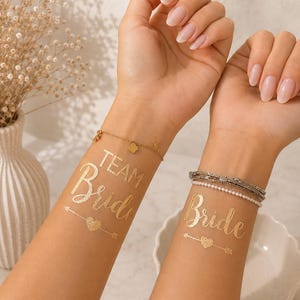 Könnte beinhalten: Zwei metallisch goldene temporäre Tattoos auf den Innenseiten der Handgelenke. Ein Tattoo zeigt den Schriftzug "TEAM Bride" über einem Herz- und Pfeildesign. Das andere Tattoo zeigt den Schriftzug "Bride" über einem Herz- und Pfeildesign in Schreibschrift.