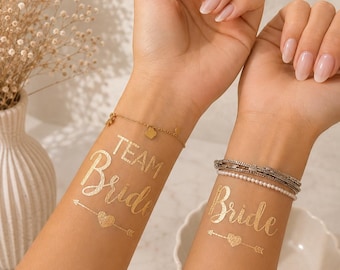 Op de afbeelding: Twee metallic gouden tijdelijke tatoeages op de binnenkant van de polsen. De ene tatoeage toont de tekst "TEAM Bride" boven een hart- en pijldesign. De andere tatoeage toont de tekst "Bride" boven een hart- en pijldesign in een sierlijk lettertype.
