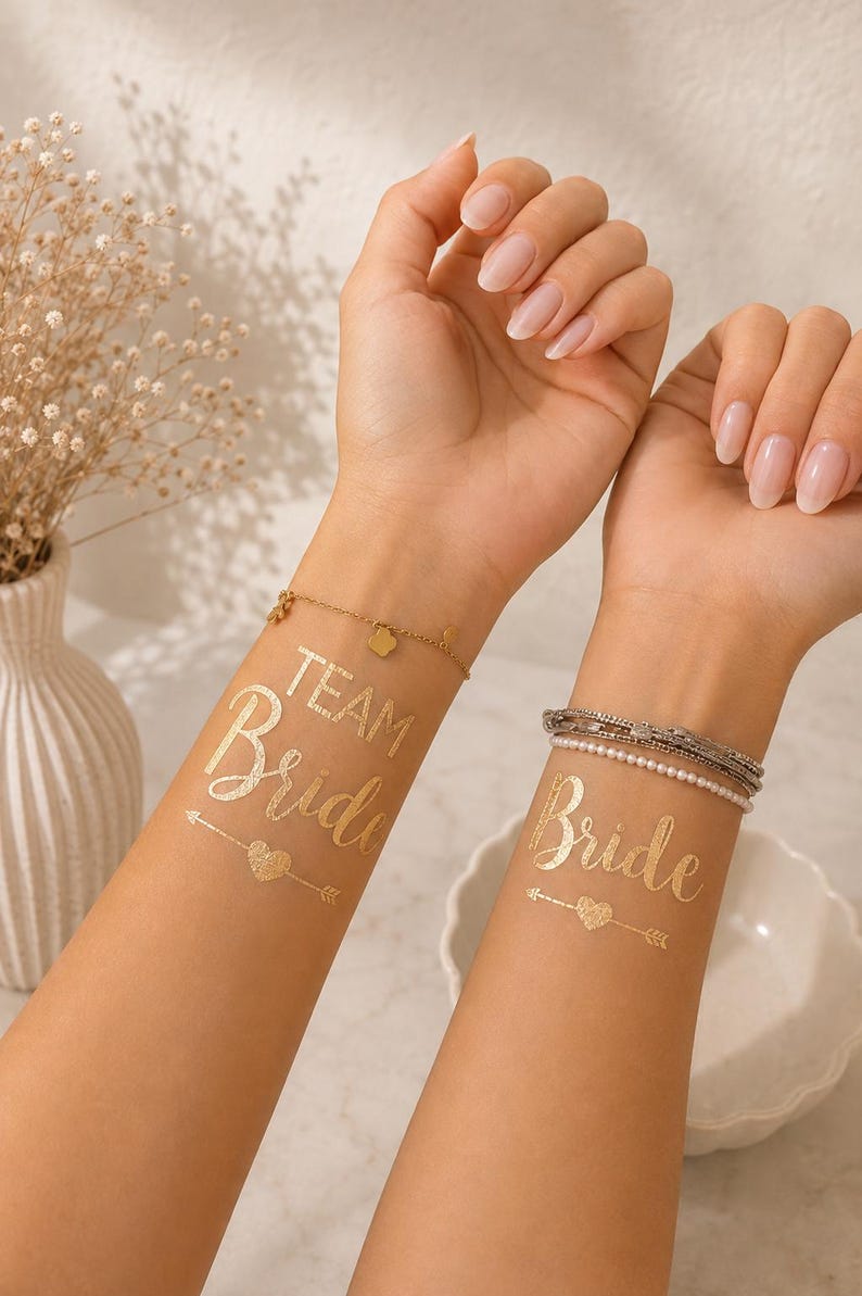 Op de afbeelding: Twee metallic gouden tijdelijke tatoeages op de binnenkant van de polsen. De ene tatoeage toont de tekst "TEAM Bride" boven een hart- en pijldesign. De andere tatoeage toont de tekst "Bride" boven een hart- en pijldesign in een sierlijk lettertype.