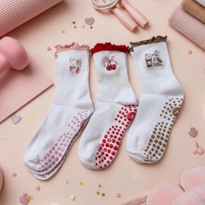 Könnte beinhalten: Drei Paar weiße Socken mit farbigen Verzierungen und Designs. Ein Paar hat rosa Verzierungen und ein Schleifenmuster, ein anderes hat rote Verzierungen und Kirschen, und das dritte hat braune Verzierungen und eine Kaffeetasse. Die Sohlen haben Anti-Rutsch-Griffe.