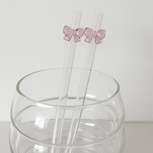 Op de afbeelding: Twee heldere glazen rietjes met roze strikaccenten staan in een heldere glazen kom. De rietjes zijn recht en hebben een kleine, decoratieve strik aan de bovenkant. De kom is rond en leeg.