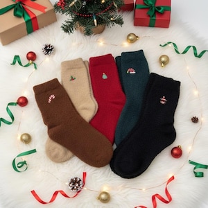 Peut inclure: Cinq paires de chaussettes de Noël en marron, beige, rouge, vert foncé et noir. Chaque chaussette est ornée d'une broderie festive. Les chaussettes sont présentées sur une surface blanche et duveteuse, entourées de décorations de Noël.