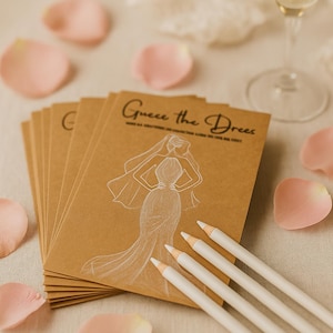 Op de afbeelding: Kraftpapierkaarten met de tekst "Guess the Dress" en een bruidsillustratie, samen met witte potloden en roze rozenblaadjes. Op de achtergrond is een champagneglas te zien, wat wijst op een spel voor een bruidsfeest.