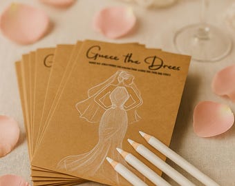 Op de afbeelding: Kraftpapierkaarten met de tekst "Guess the Dress" en een bruidsillustratie, samen met witte potloden en roze rozenblaadjes. Op de achtergrond is een champagneglas te zien, wat wijst op een spel voor een bruidsfeest.