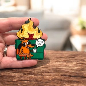 Könnte beinhalten: Ein origineller Ansteck-Pin mit einem orangefarbenen Hund, der in einer grünen Box mit Flammen sitzt. Der Hund hält einen weißen Becher und der Text "THIS IS FINE" steht in einer Sprechblase. Die Flammen haben goldenen Glitzer.