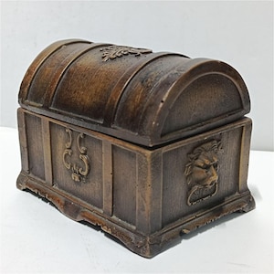 Op de afbeelding: Een bruine, rechthoekige opbergdoos met een gebogen, scharnierend deksel. De buitenkant heeft een houtnerftextuur, een decoratief krulmotief op de bovenkant en een reliëf van een leeuwenkop met een ring op het zijpaneel.