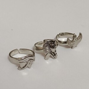 May include: Three silver-colored adjustable rings with Egyptian-themed designs. One ring features the Eye of Horus, another a pharaoh's head, and the third an eagle with outstretched wings. The rings are displayed on a white surface.