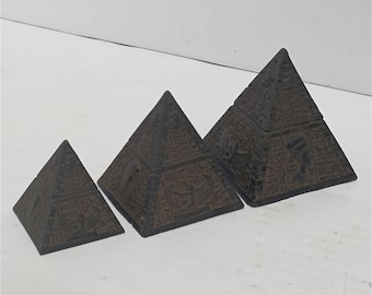 Könnte beinhalten: Drei dunkelbraune pyramidenförmige Boxen in verschiedenen Größen. Jede Pyramide hat detaillierte Schnitzereien ägyptischer Hieroglyphen und Figuren. Die Boxen sind in einer Reihe angeordnet und zeigen ihre komplizierten Designs.