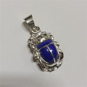 May include: A silver pendant shaped like a scarab beetle, featuring a smooth, deep blue oval stone set into the center of the metallic body. The pendant includes a polished silver bail at the top for attachment to a chain or cord necklace.