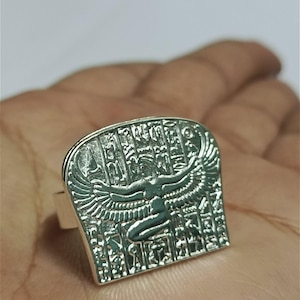 May include: Silver-toned ring featuring an Egyptian goddess design with outstretched wings and hieroglyphs. The ring has a rectangular face with intricate details, and is held in a hand.