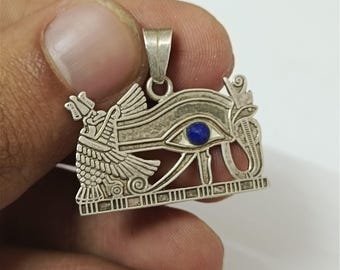 May include: Silver pendant featuring the Eye of Horus, an ancient Egyptian symbol. The pendant has intricate details, including a blue stone representing the eye's pupil. The pendant is suspended from a small loop.