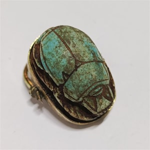 May include: A brooch featuring a carved turquoise-colored scarab beetle with dark brown weathered detailing. The scarab is mounted in a polished gold-tone metal setting with a visible pin mechanism on the side, showcasing an oval shape.