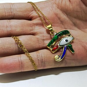 May include: Gold-colored chain necklace with a pendant featuring the Eye of Horus. The pendant is detailed with green, red, white, and blue enamel. The necklace is displayed on a hand.