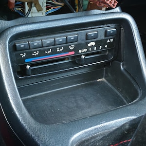 Puede incluir: Un panel de control de climatización automotriz negro con palancas deslizantes horizontales para la temperatura y la velocidad del ventilador, botones para aire acondicionado y funciones de desempañado, y un compartimento de almacenamiento.