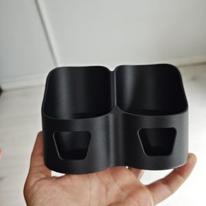 Op de afbeelding: Een matzwarte organizer met twee compartimenten, voorzien van twee diepe, afgeronde rechthoekige opbergvakken en twee kleine, trapeziumvormige uitsparingen aan de voorzijde. De structuur is gemaakt van een stevig materiaal met een textuur.