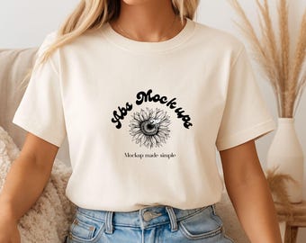 May include: Cream-colored t-shirt with a black sunflower graphic and the text "Abs Mockups" and "Mockup made simple". The shirt has short sleeves and a crew neck. The model is wearing blue jeans.