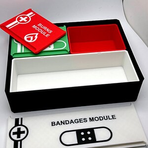 May include: A black first aid kit with red, green, and white compartments. The kit includes a red lid labelled "BURNS MODULE" and a white lid labelled "BANDAGES MODULE". The kit is designed for organising medical supplies.
