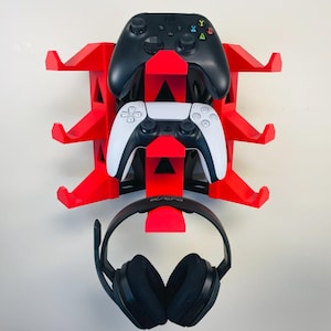 May include: A red wall-mounted gaming controller and headset holder. The holder has three levels, with a black Xbox controller on top, a white and black PlayStation controller in the middle, and a black headset at the bottom.