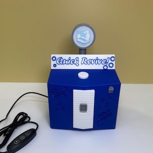 Może przedstawiać: Niebiesko-białe urządzenie z napisem "Quick Revive!" i okrągłym znakiem na górze. Urządzenie ma biały panel przedni z przyciskiem oraz napisami "Ice Cold" i "Sold Here" po bokach. Podłączony jest czarny kabel i panel sterowania.