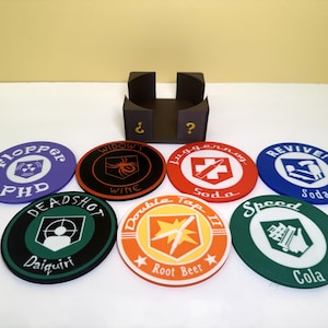 Peut inclure: Ensemble de huit dessous de verre circulaires colorés avec divers motifs et textes, dont "Flopper PHD", "Widow's Wine" et "Juggernog Soda". Un porte-gobelet noir avec des points d'interrogation est également visible.