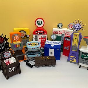 Op de afbeelding: Een verzameling kleurrijke, miniatuur arcade-achtige machines, waaronder een bruine automaat met "5¢" op de voorkant, een rood-witte frisdrankautomaat en een paarse machine met een geel paneel. De machines hebben verschillende logo's en tekst.