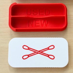 Puede incluir: Un organizador de plástico rojo con las palabras "USED" y "NEW" impresas, y una tarjeta blanca con un gráfico de bastoncillos de algodón rojos. El organizador tiene dos compartimentos.