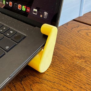 May include: A yellow laptop stand supports a black laptop on a wooden surface. The stand has a curved, hook-shaped design, providing elevation. The laptop screen displays various application icons.