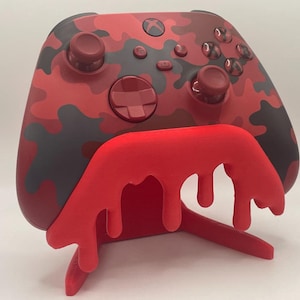 May include: A red and black camouflage Xbox controller resting on a matching red stand. The stand has a unique dripping design. The controller features buttons and joysticks in red.